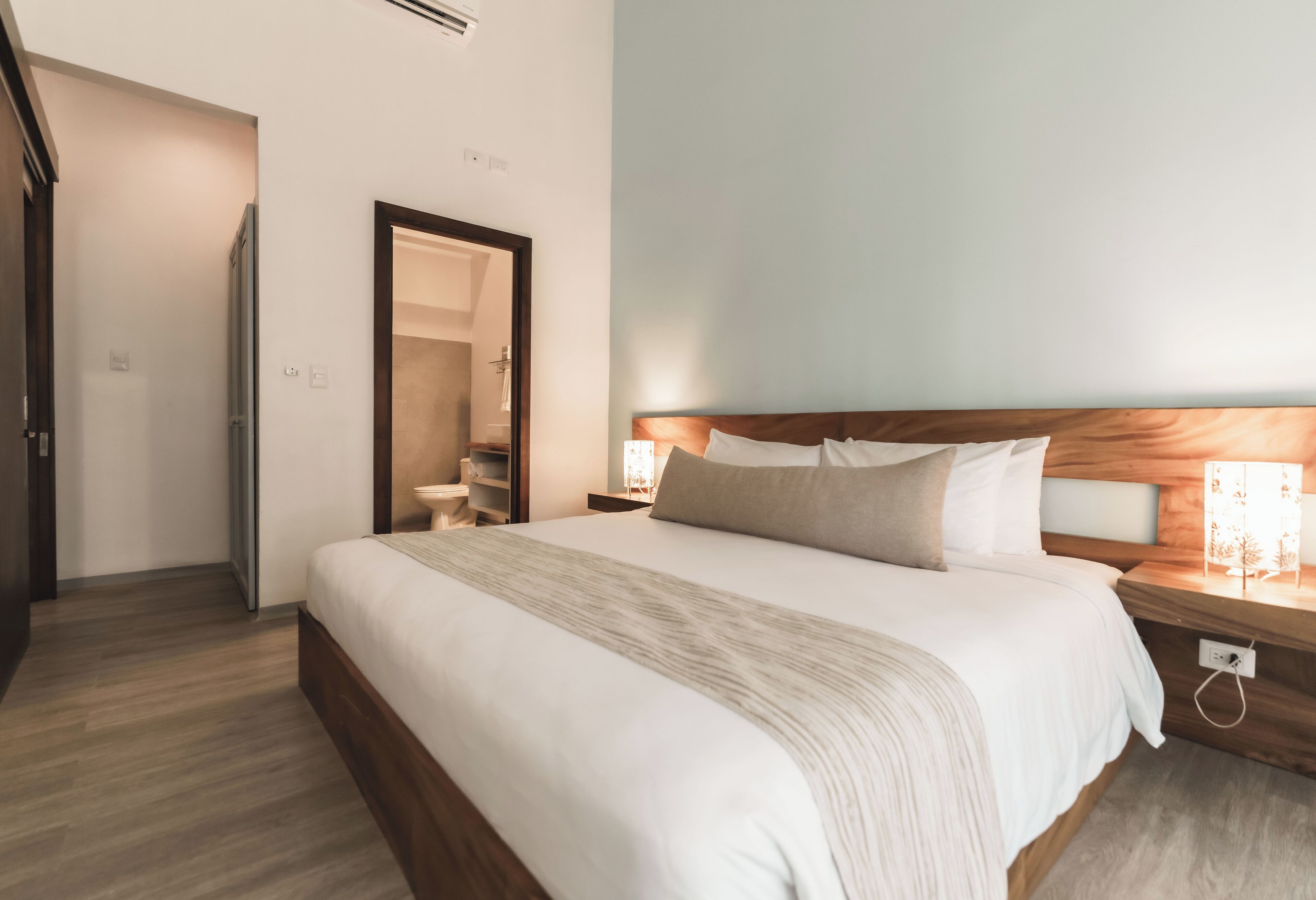 Ein ordentlich eingerichtetes Hotelzimmer mit einem großen Bett, Nachttischlampen, einem Holzkopfende und einem Spiegel.