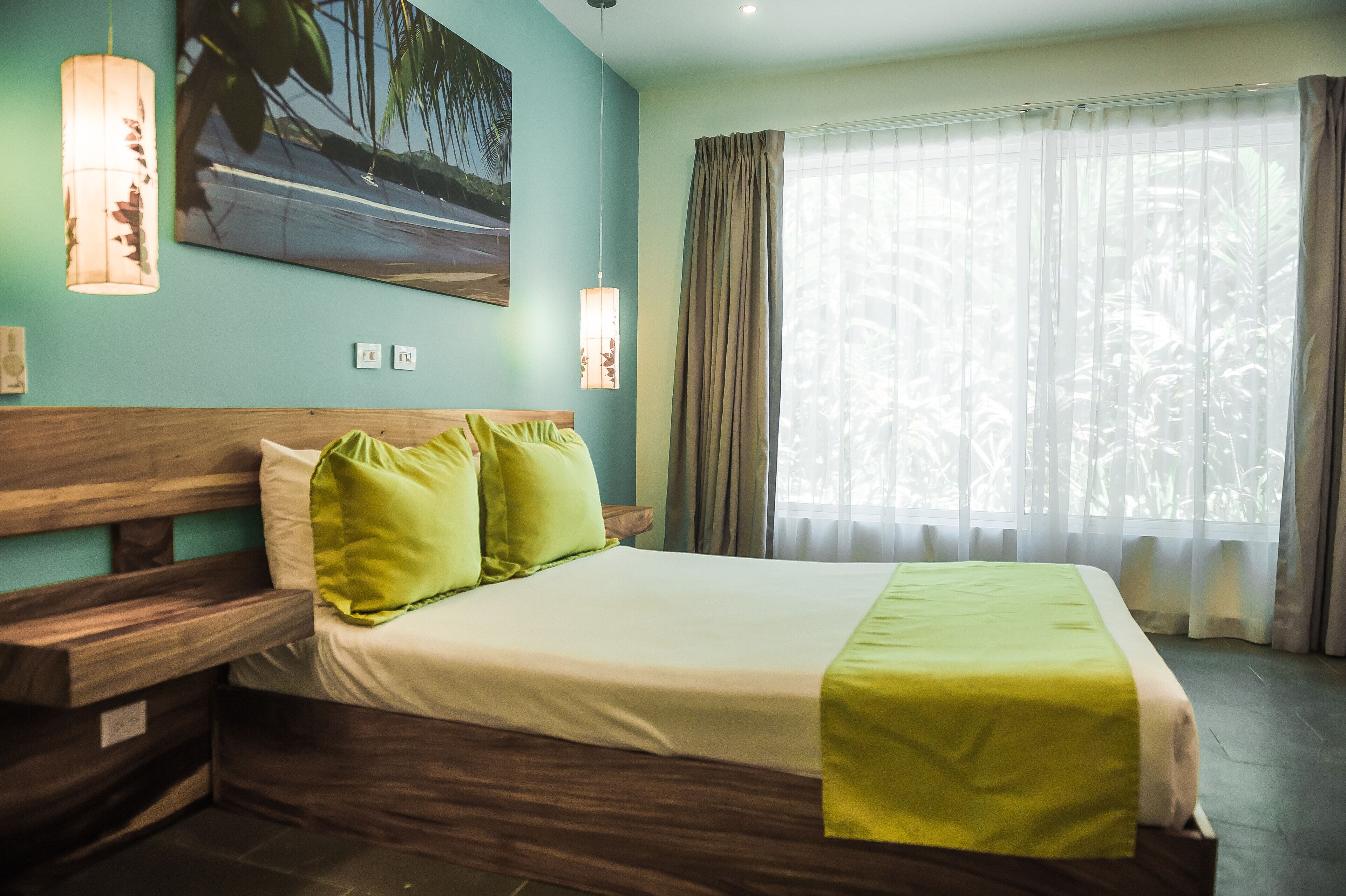 Una cama bien hecha con una manta verde, un cabecero de madera y dos almohadas verdes. En la pared hay un cuadro de un camino, y una ventana con cortinas translúcidas.