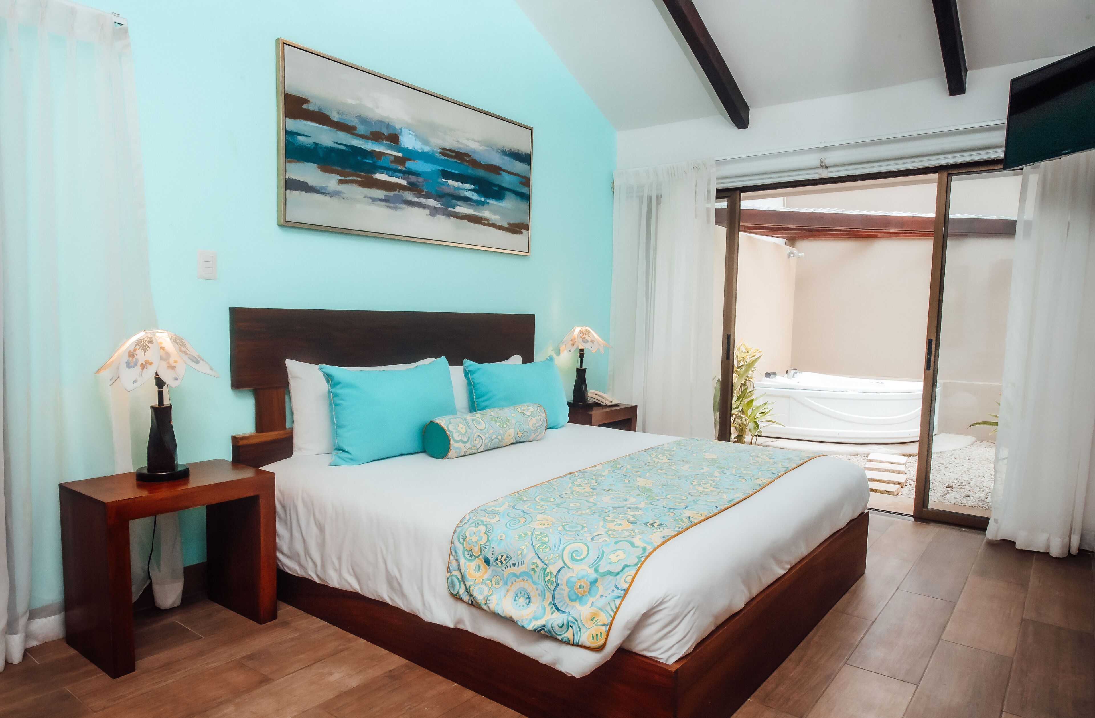 A bedroom with a bed, nightstands, a painting, and a view of a garden through a sliding door.