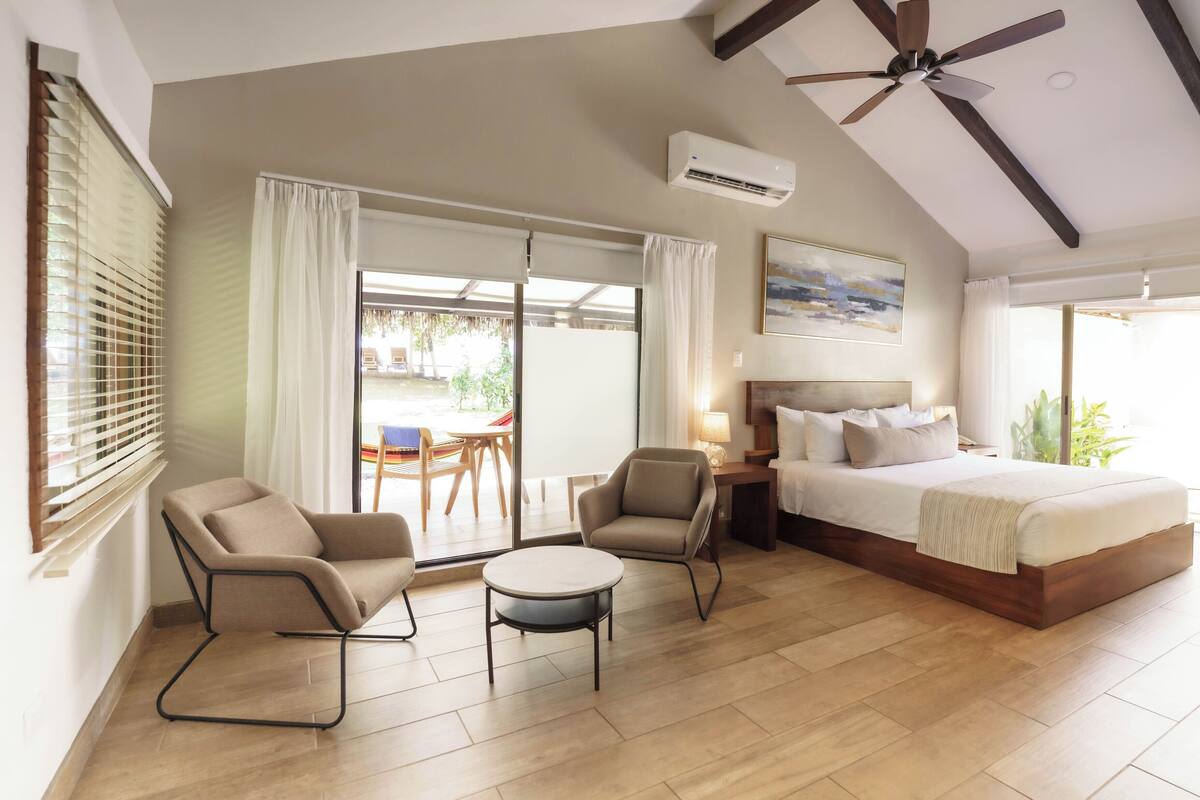 Une chambre à coucher comprenant un lit, deux chaises, une petite table, un ventilateur de plafond et une grande fenêtre avec des rideaux.