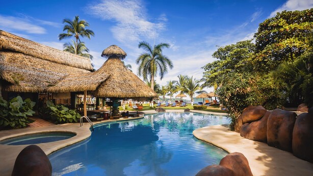 Een zwembad bij een resort met overkappingen van riet, palmbomen en een helderblauwe lucht.
