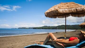 A beach scene with a person lying on a lounge chair under a straw umbrella.