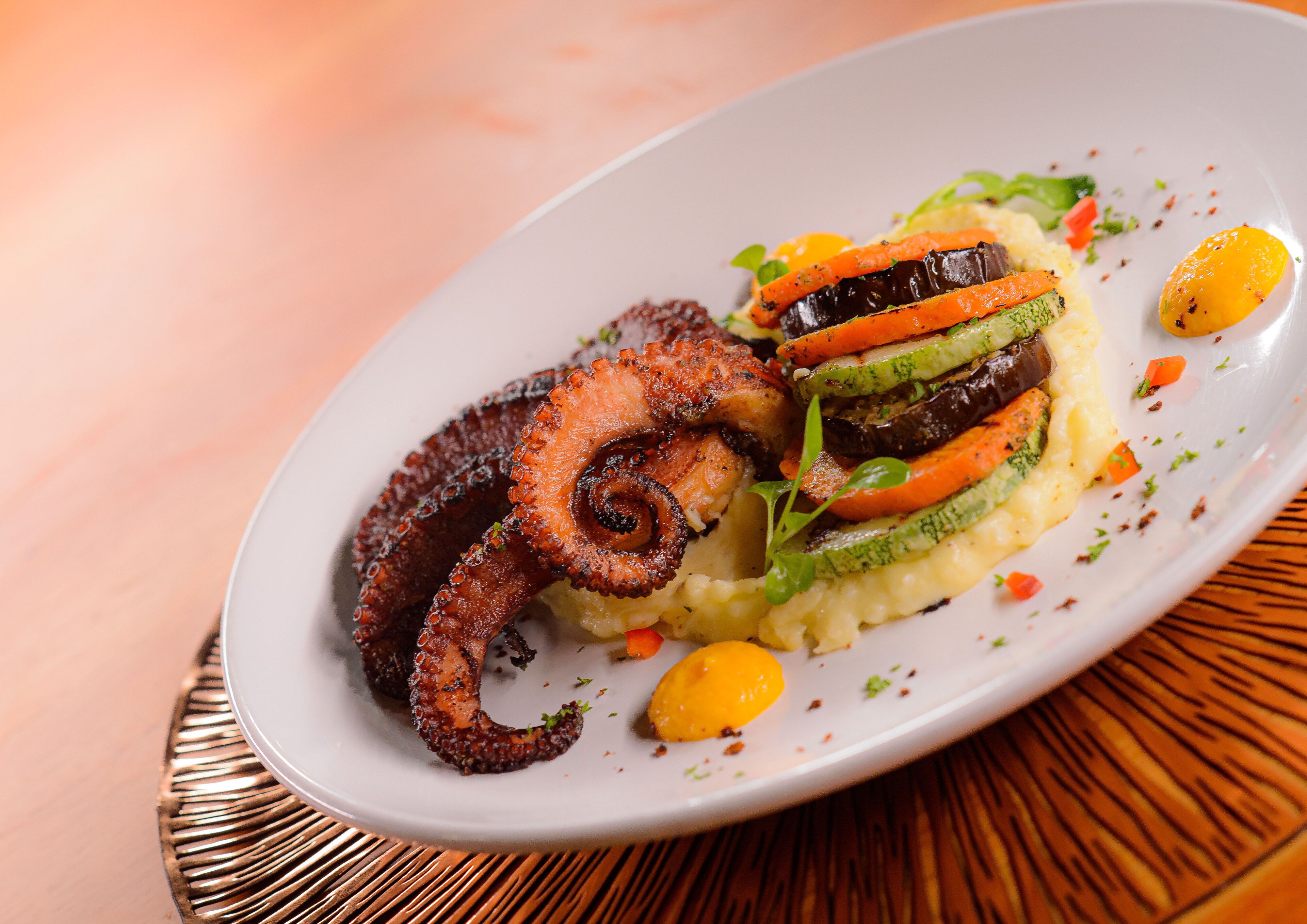 Une assiette de nourriture comprenant du poulpe, des légumes et une garniture, le tout présenté sur une assiette blanche.