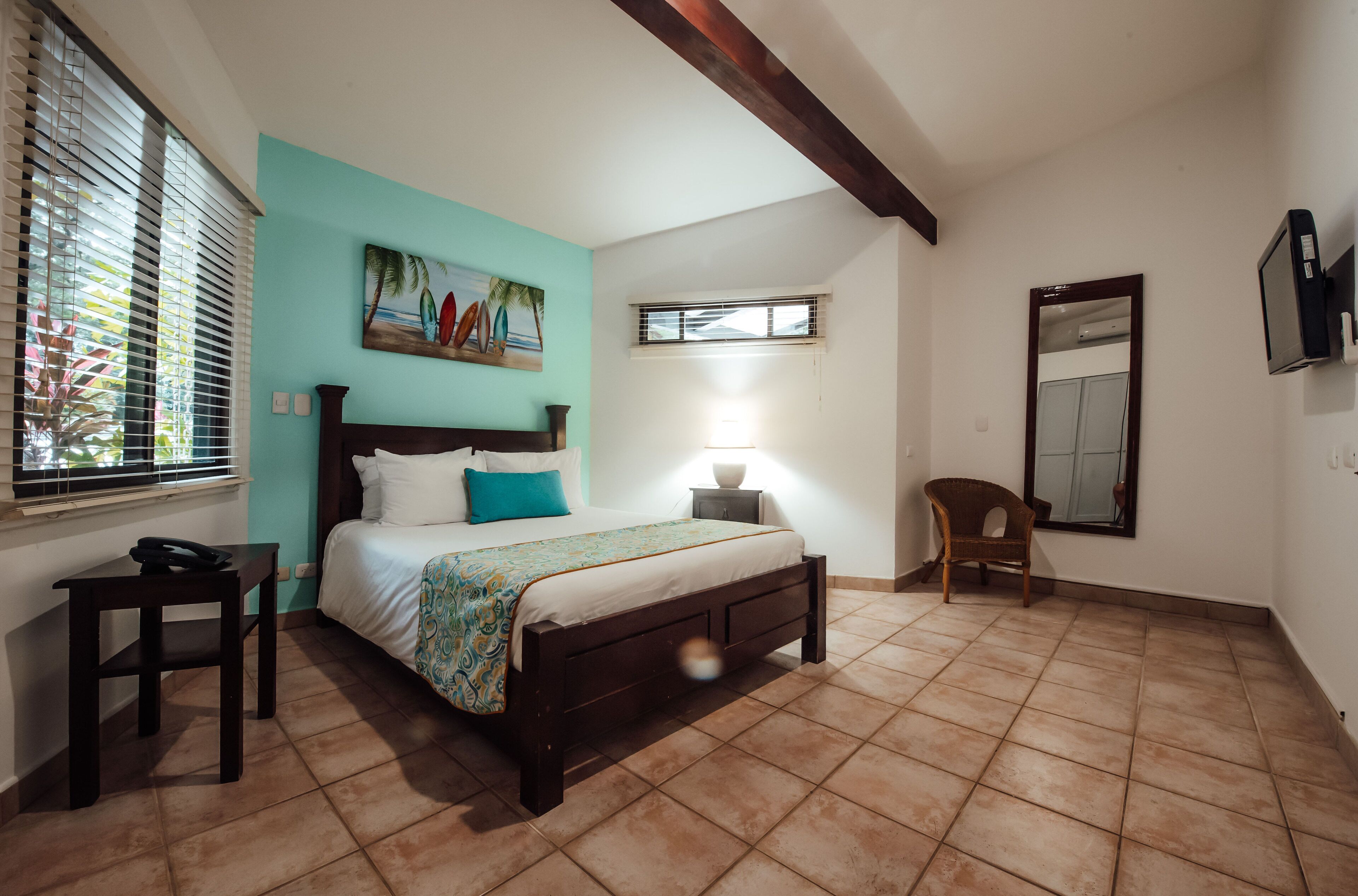 Un dormitorio con cama, mesita de noche, silla, ventana con persianas, un cuadro, televisor en la pared y ventilador de techo.