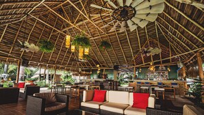 Een ruim buitengebied met rieten meubels en rode kussens, een bar met rieten dak en een grote ventilator aan het plafond.