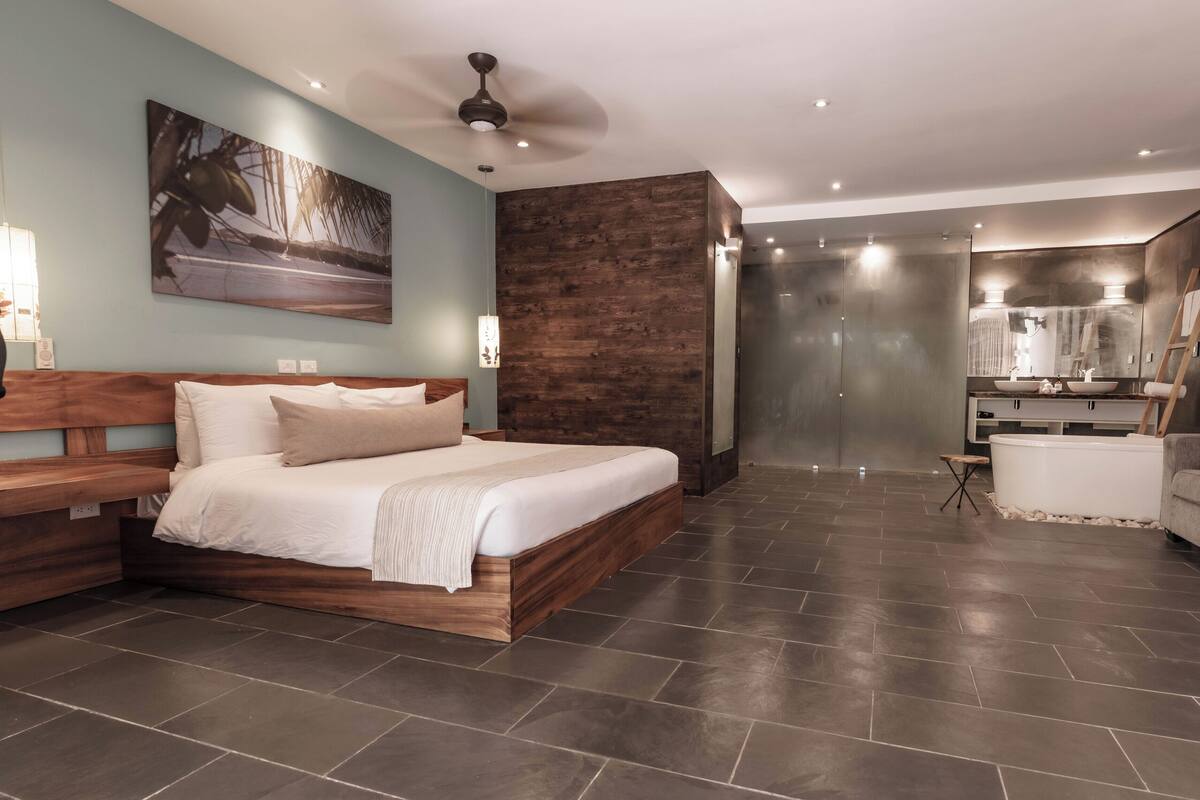 Une chambre moderne avec un grand lit, une tête de lit en bois, un ventilateur de plafond et une salle de bain dotée d’un grand lavabo et d’un grand miroir.
