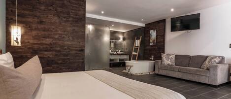 Un dormitorio moderno con una cama grande, un sofá gris, una mesita pequeña y un baño con bañera.