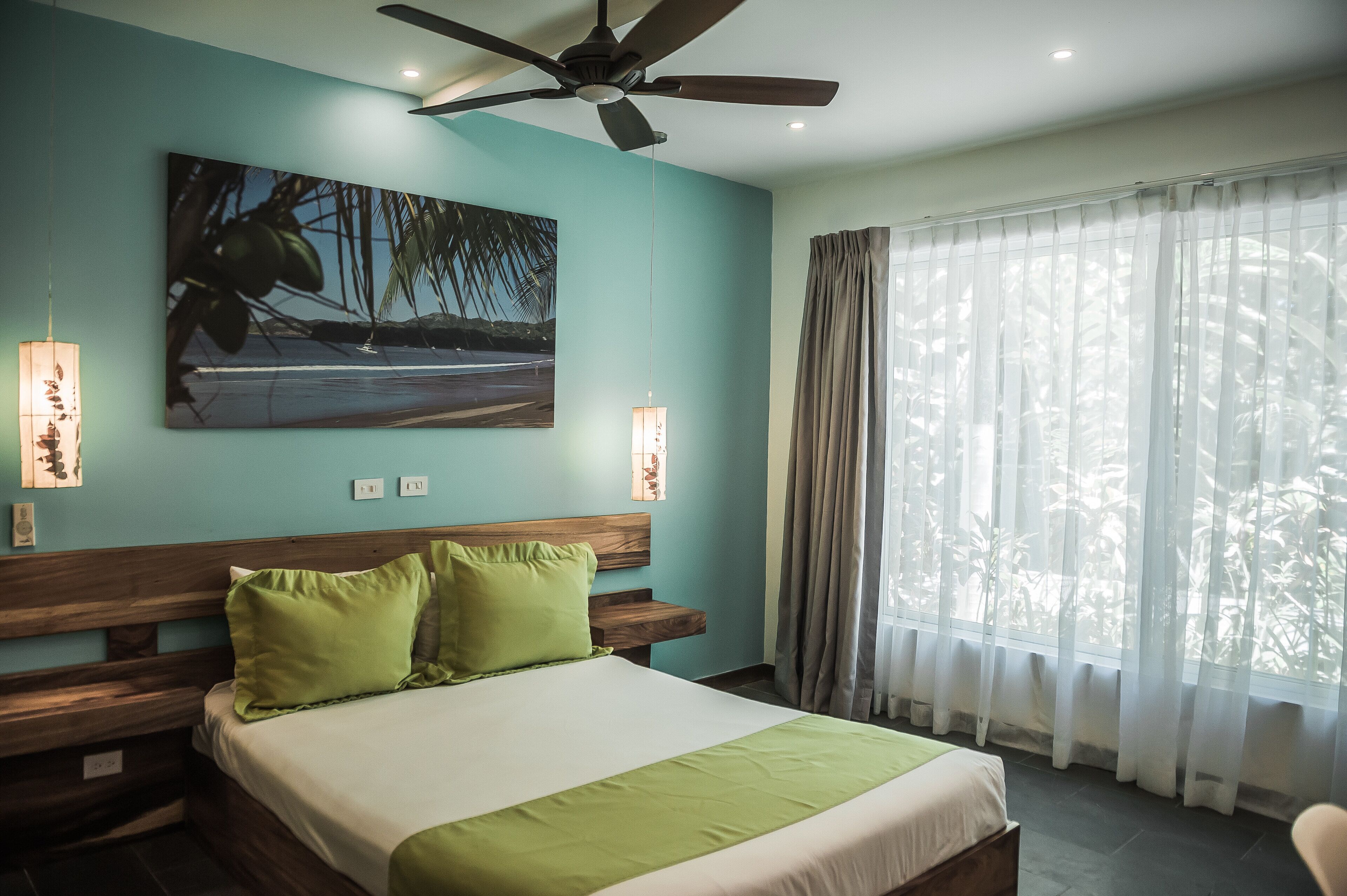 A hotel room with a bed, wooden headboard, green pillows, a ceiling fan, a painting on the wall, and a window with sheer curtains.
