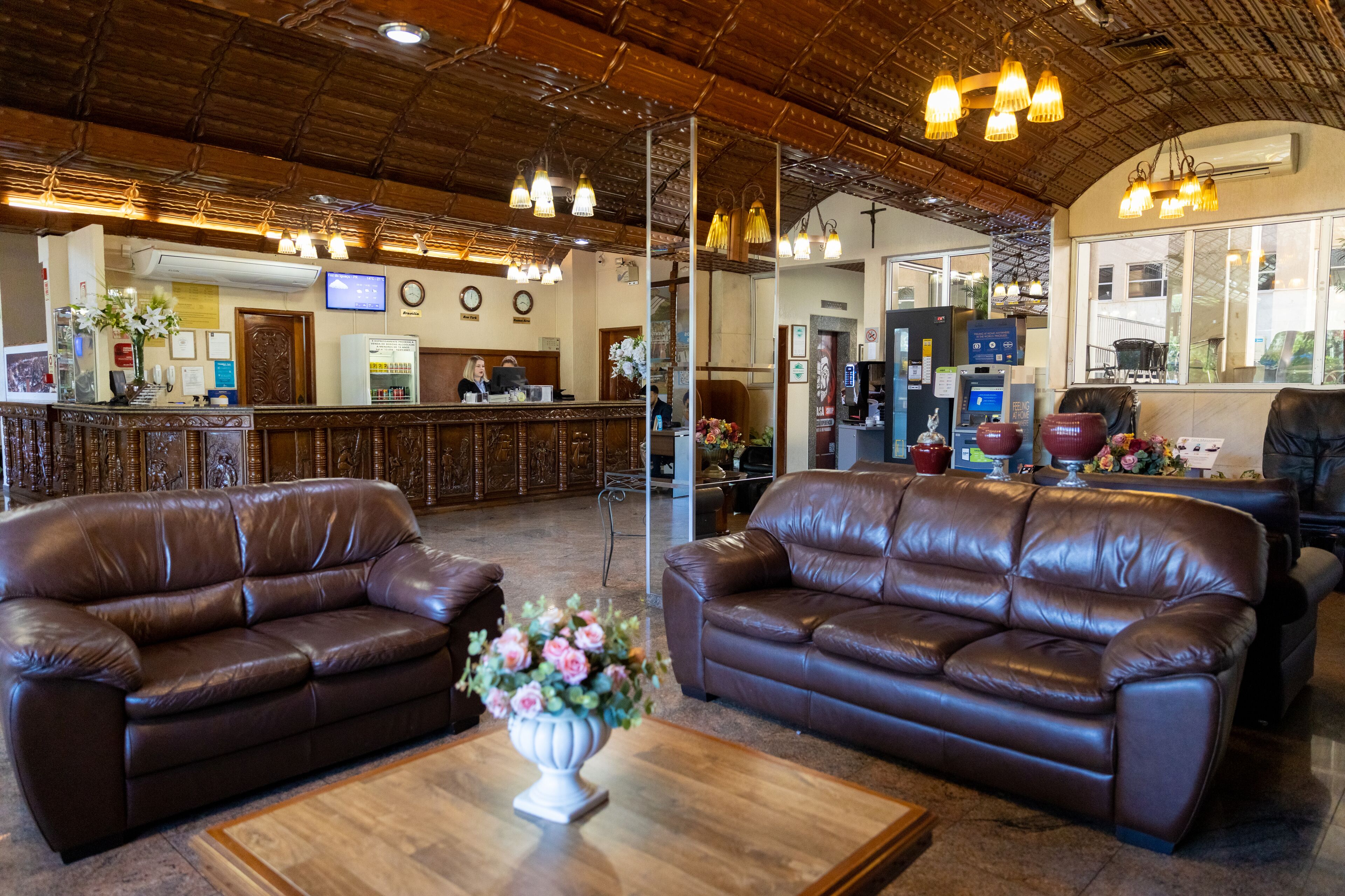 A lobby with two leather sofas, a wooden coffee table with a vase of flowers, a reception desk, and a large window with a view.