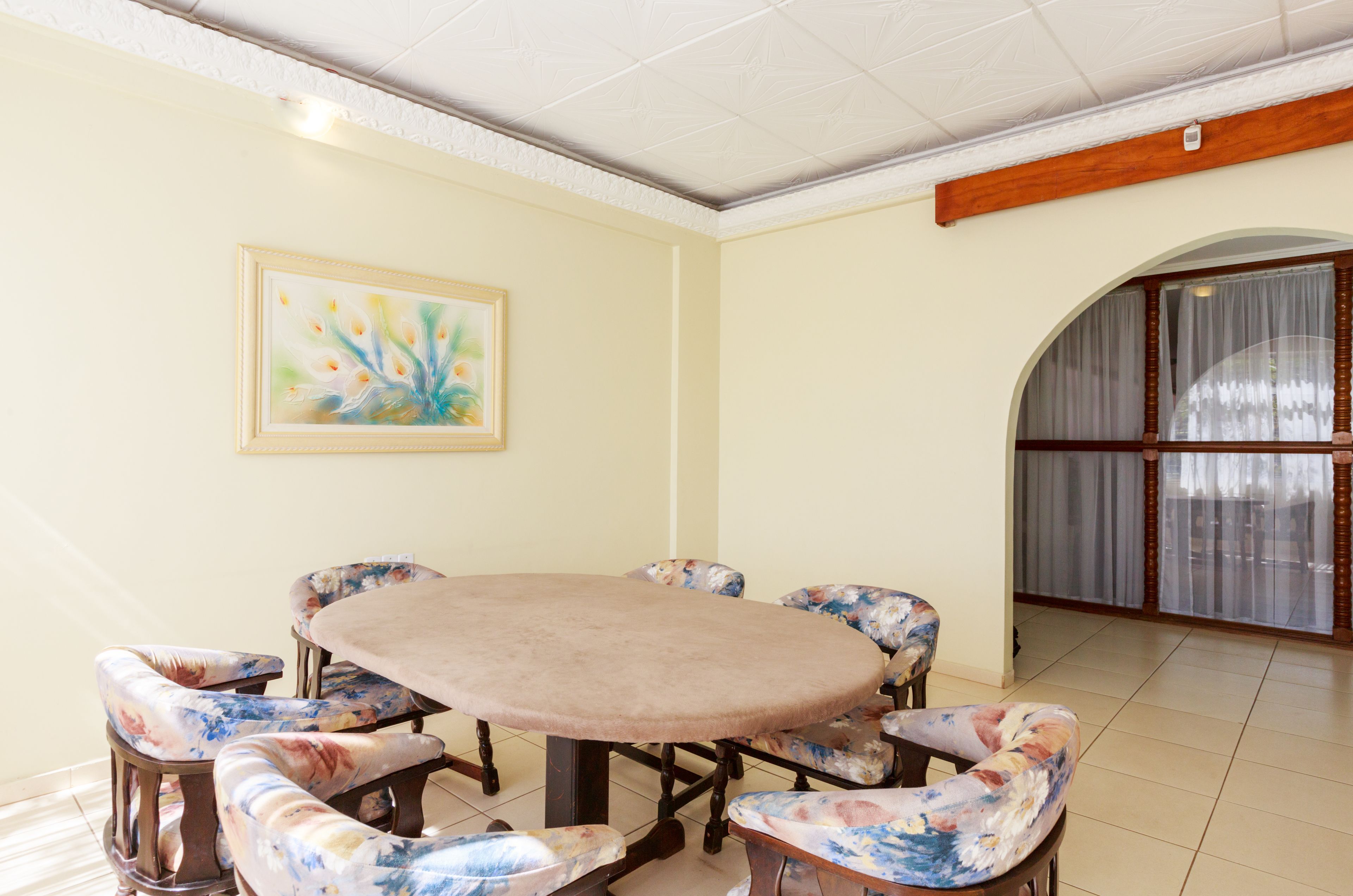 A dining area with a round table and floral upholstered chairs, a framed artwork on the wall, and a large window with curtains.