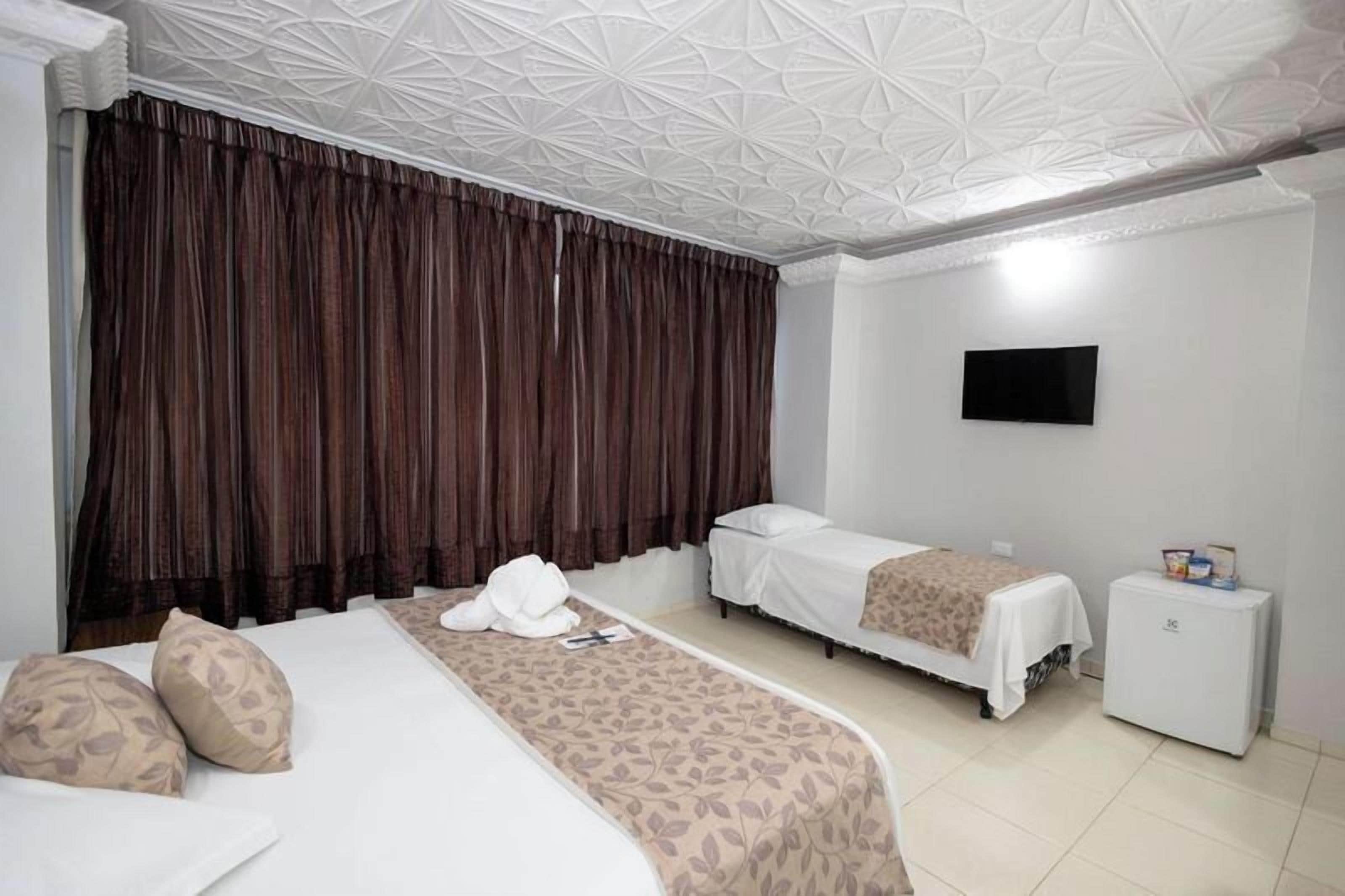 A hotel room with a large bed, two smaller beds, a flat-screen TV, a minibar, and a ceiling with a geometric pattern.