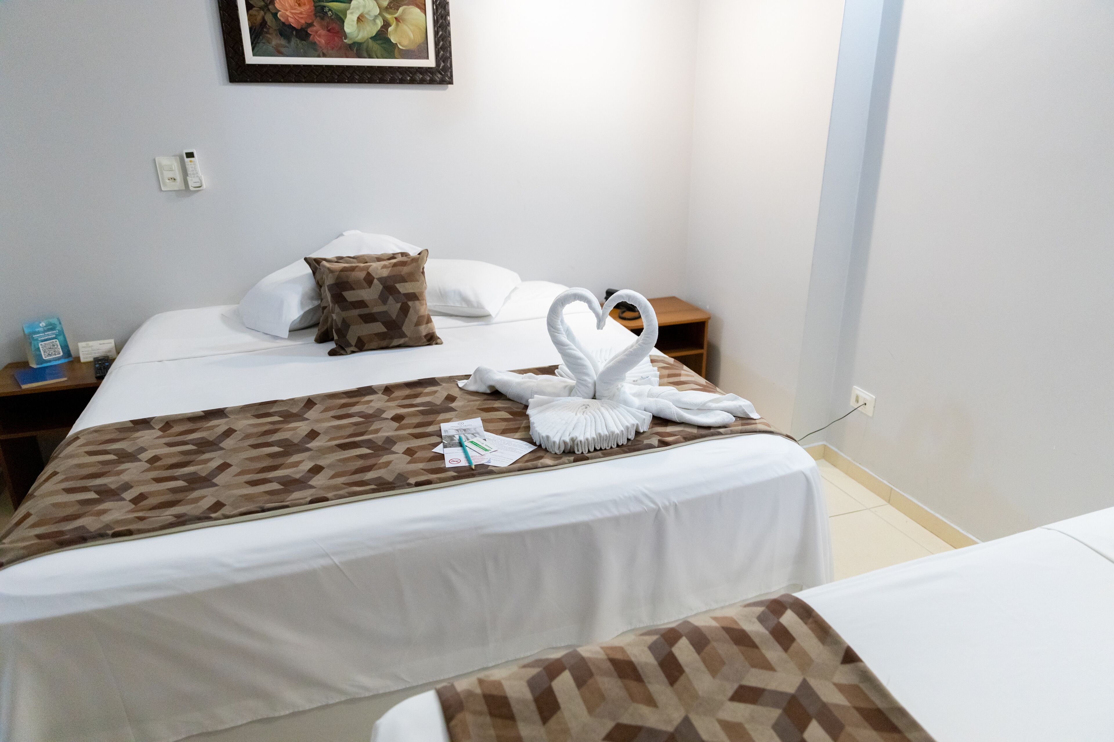 A hotel room with two beds, a patterned bedspread, a heart-shaped towel arrangement, a nightstand, and a framed picture on the wall.