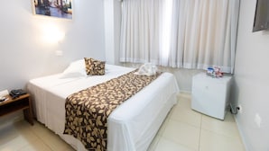 A hotel room with a bed, a bedside table, a small refrigerator, and a window with curtains.