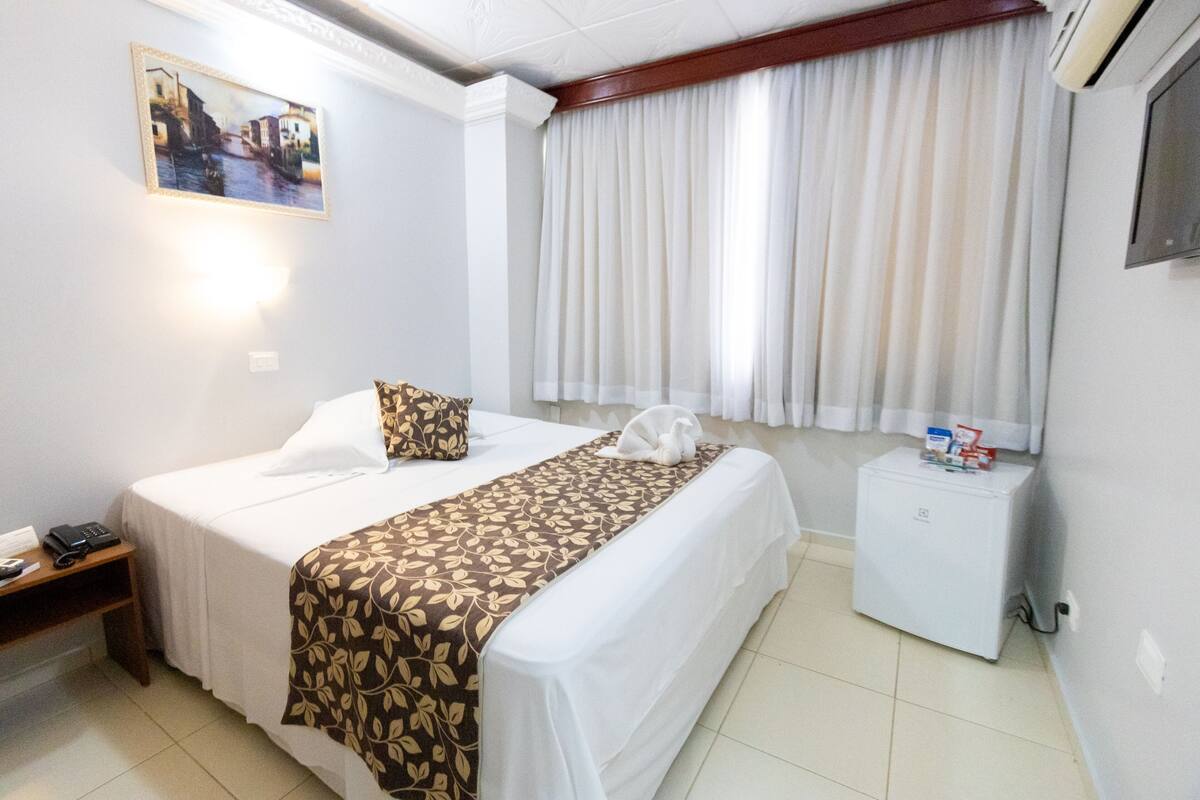 Een hotelkamer met een bed, nachtkastje, kleine koelkast en raam met gordijnen.