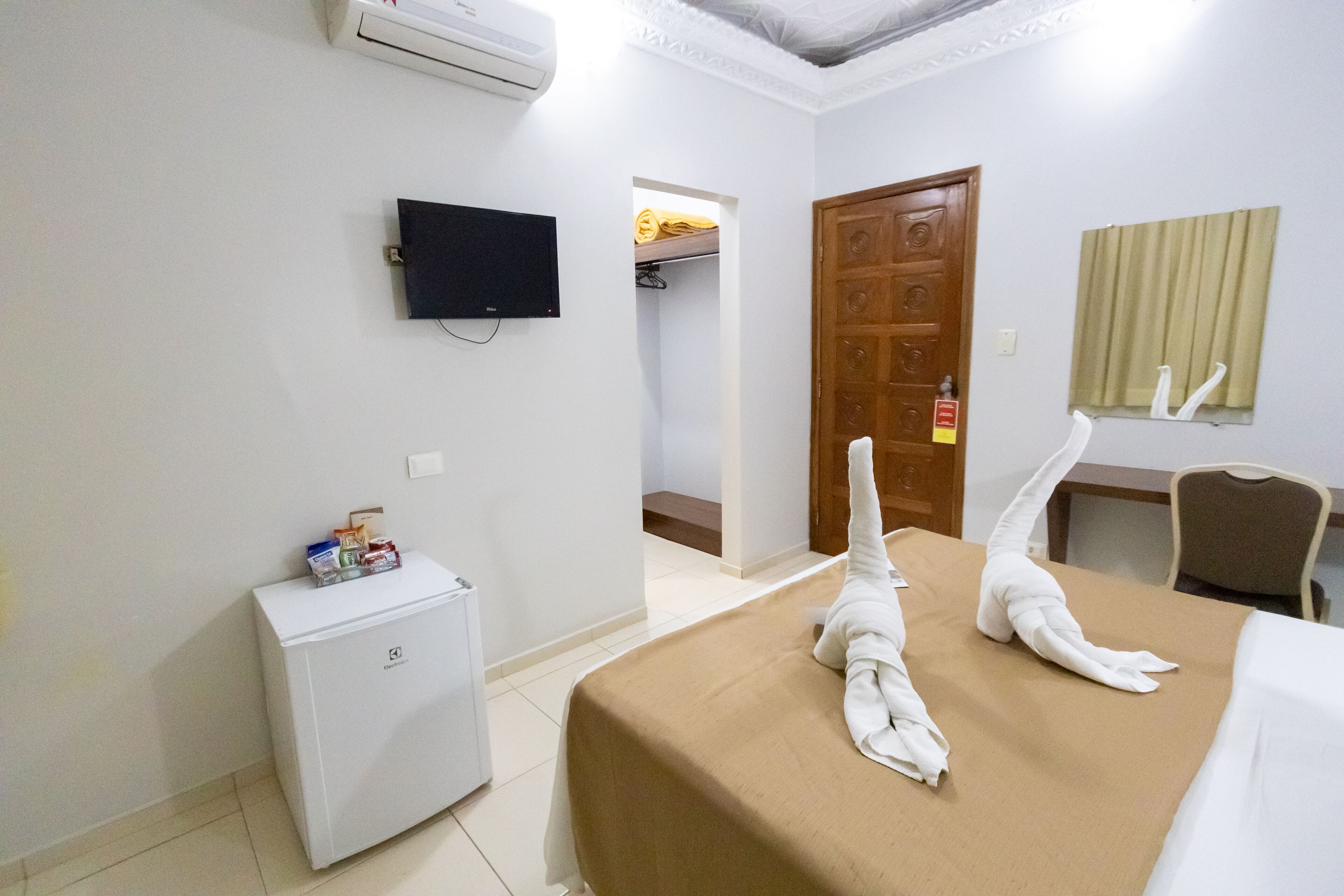 A hotel room with a bed, a small refrigerator, a television, a wooden door, and a window with curtains.