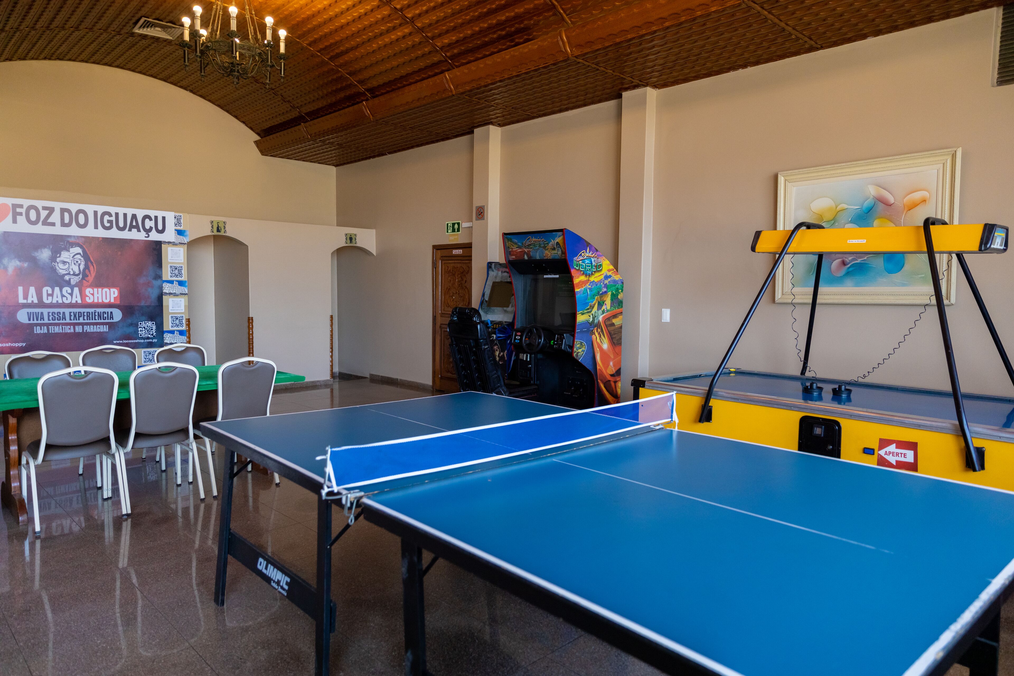A ping pong table in a room with chairs and a table, a painting on the wall, and a sign that reads "FOZ DO IGUACU".