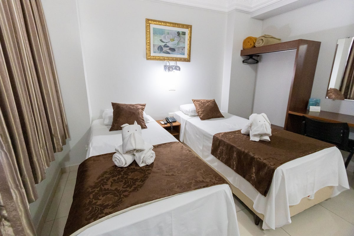 Hotelkamer met twee bedden, elk met een bruine sprei en witte lakens, een schilderij aan de muur, een spiegel en een plank met handdoeken.