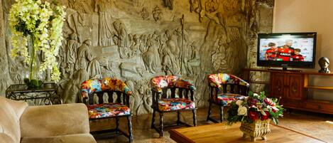 Una sala de estar con una pared de piedra, un sofá, dos sillones coloridos, una mesa de centro de madera, un televisor y un jarrón con flores.