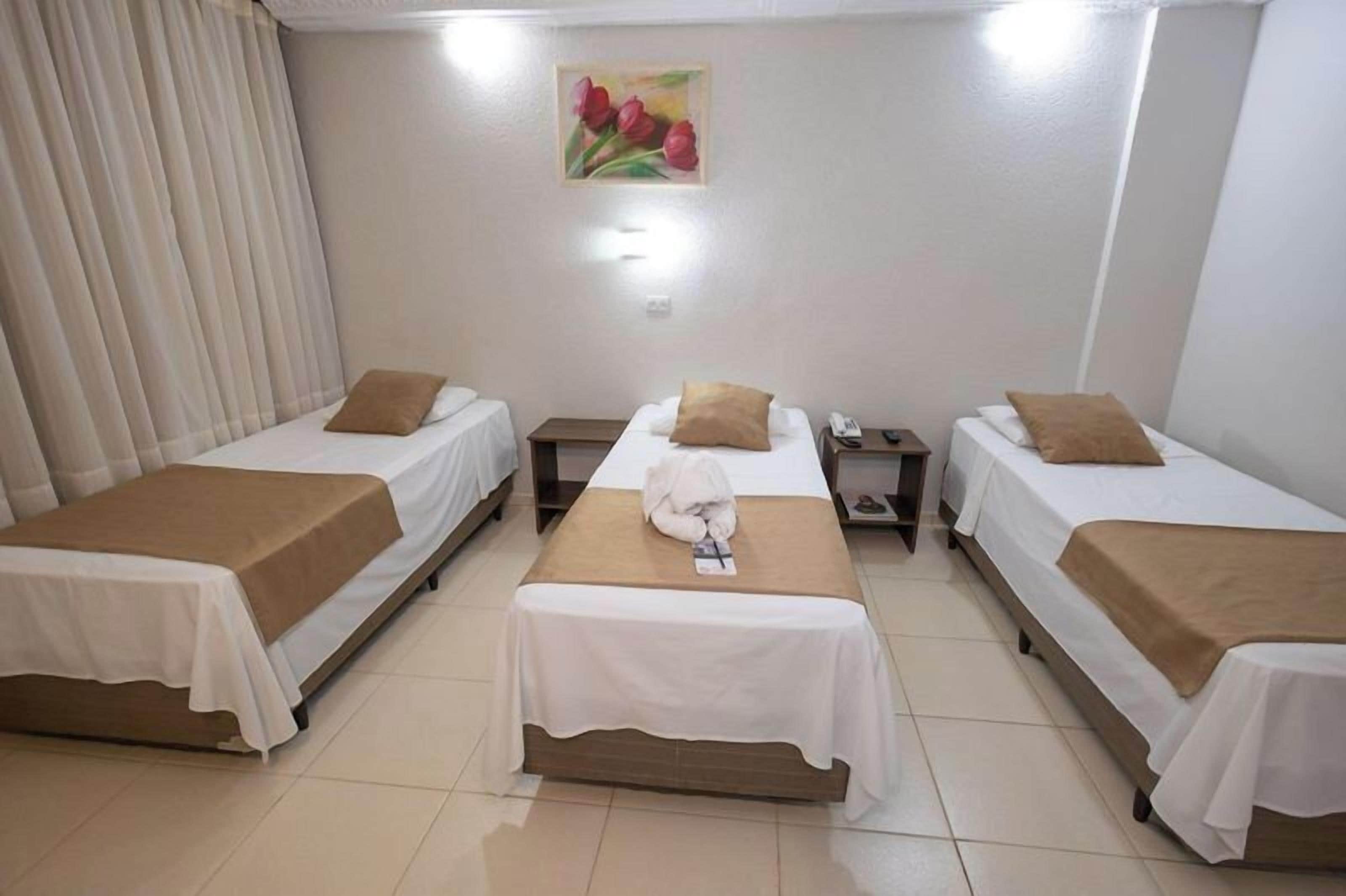 A hotel room with three single beds, each with a brown headboard and white bedding. There is a painting of red flowers on the wall above the center bed.