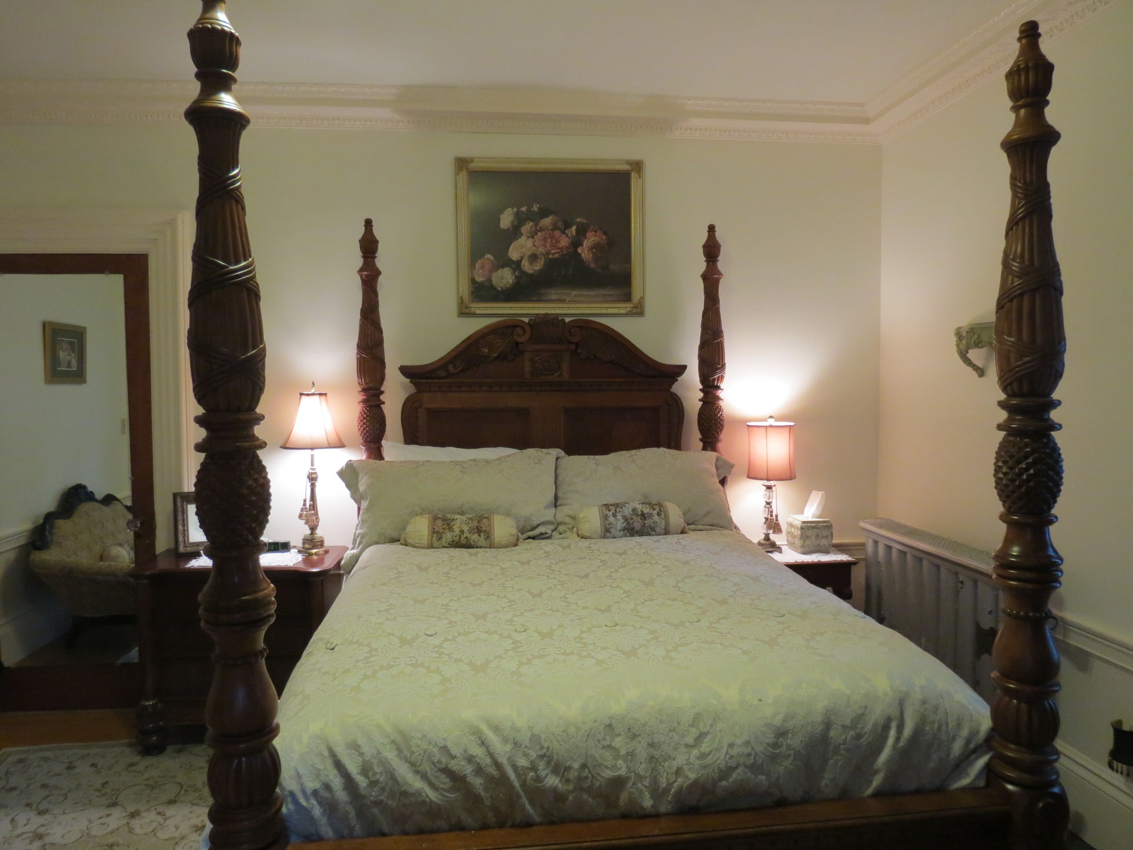 A four-poster bed with a floral headboard, two bedside lamps, and a framed painting above.