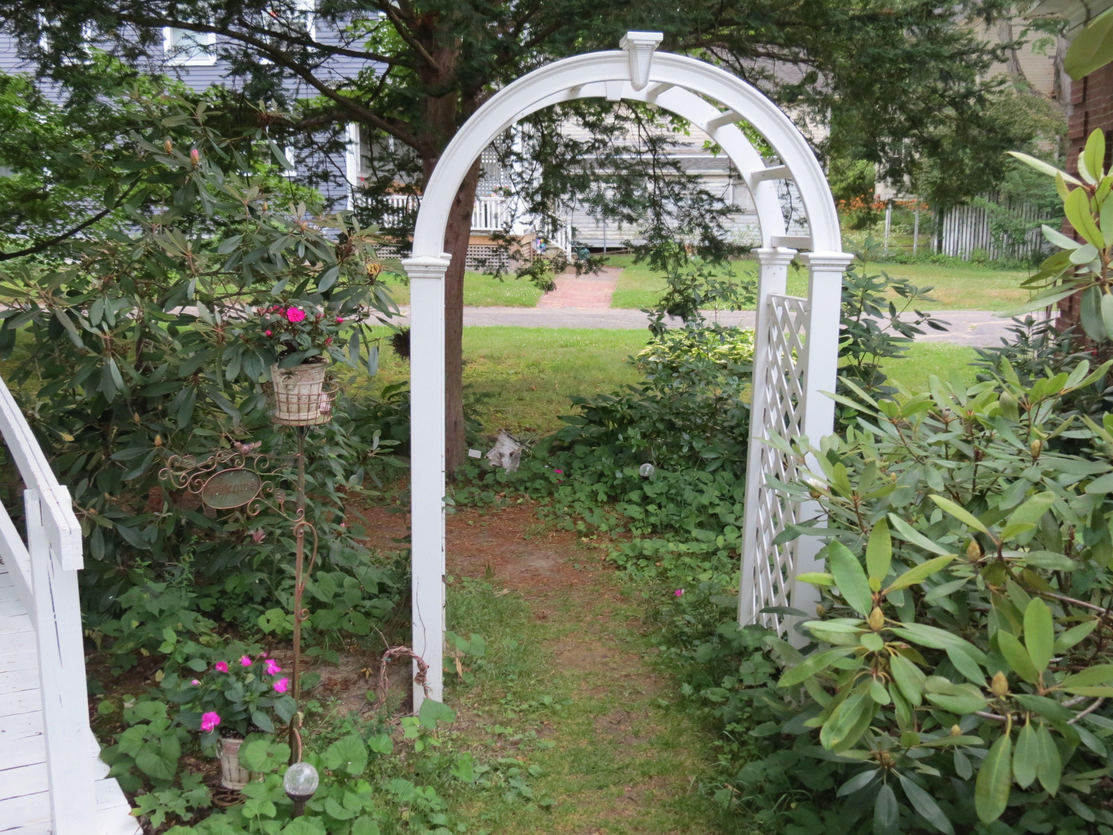 A white arbor with a pathway leading through it, surrounded by lush greenery and flowering plants.