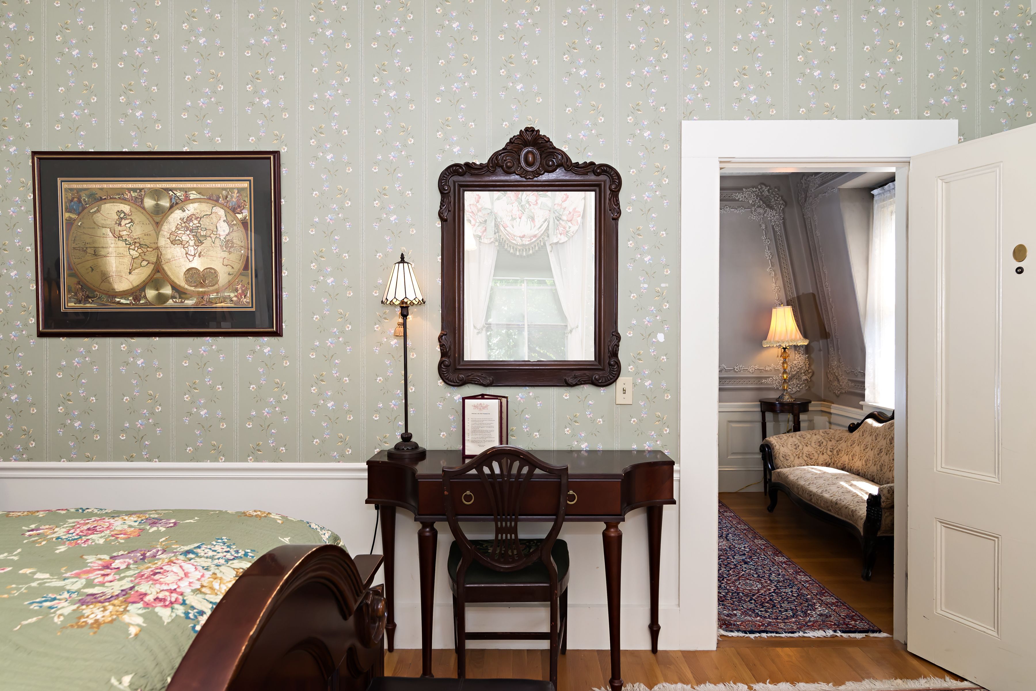 A bedroom with a bed, a desk with a chair, a dresser, a framed map, and a mirror.