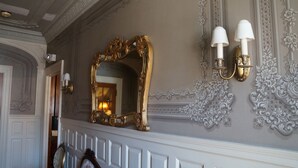 A grand hallway with an ornate gold mirror, white wall sconces, and intricate wall molding.