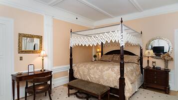 A four-poster canopy bed with a bench, a desk with a chair, a mirror, and a nightstand with a lamp.