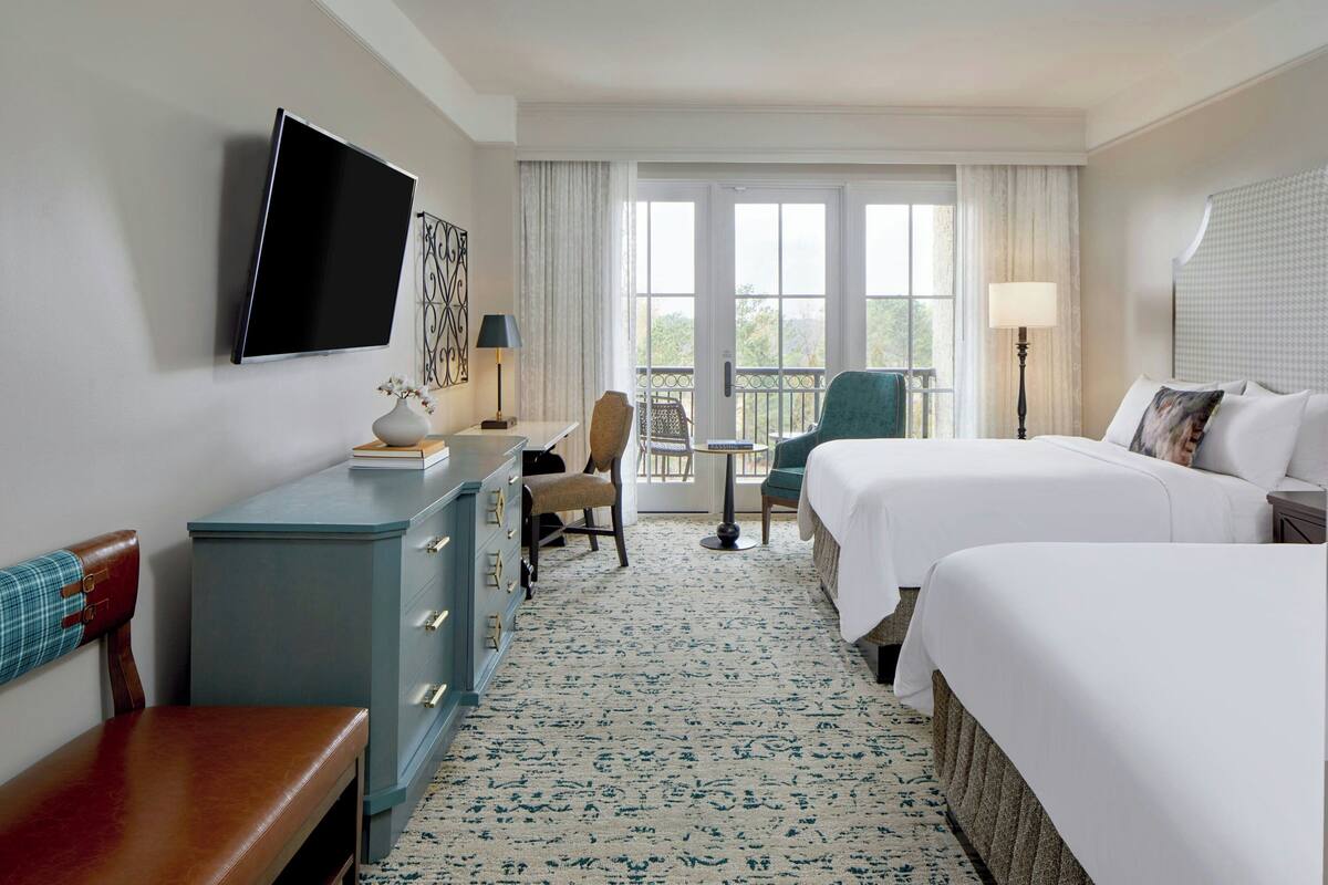 A hotel room with two beds, a desk with a chair, a TV mounted on the wall, and a balcony with a view.