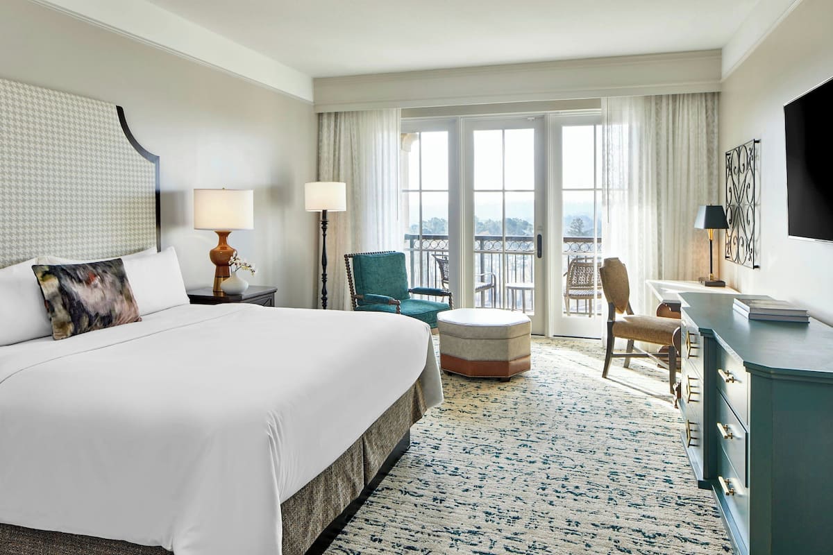 A hotel room with a large bed, a desk, a chair, a TV, and a balcony with a view.
