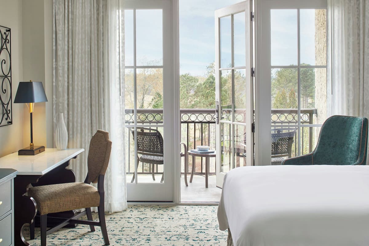A hotel room with a bed, a desk, a chair, and a balcony with a view of trees.