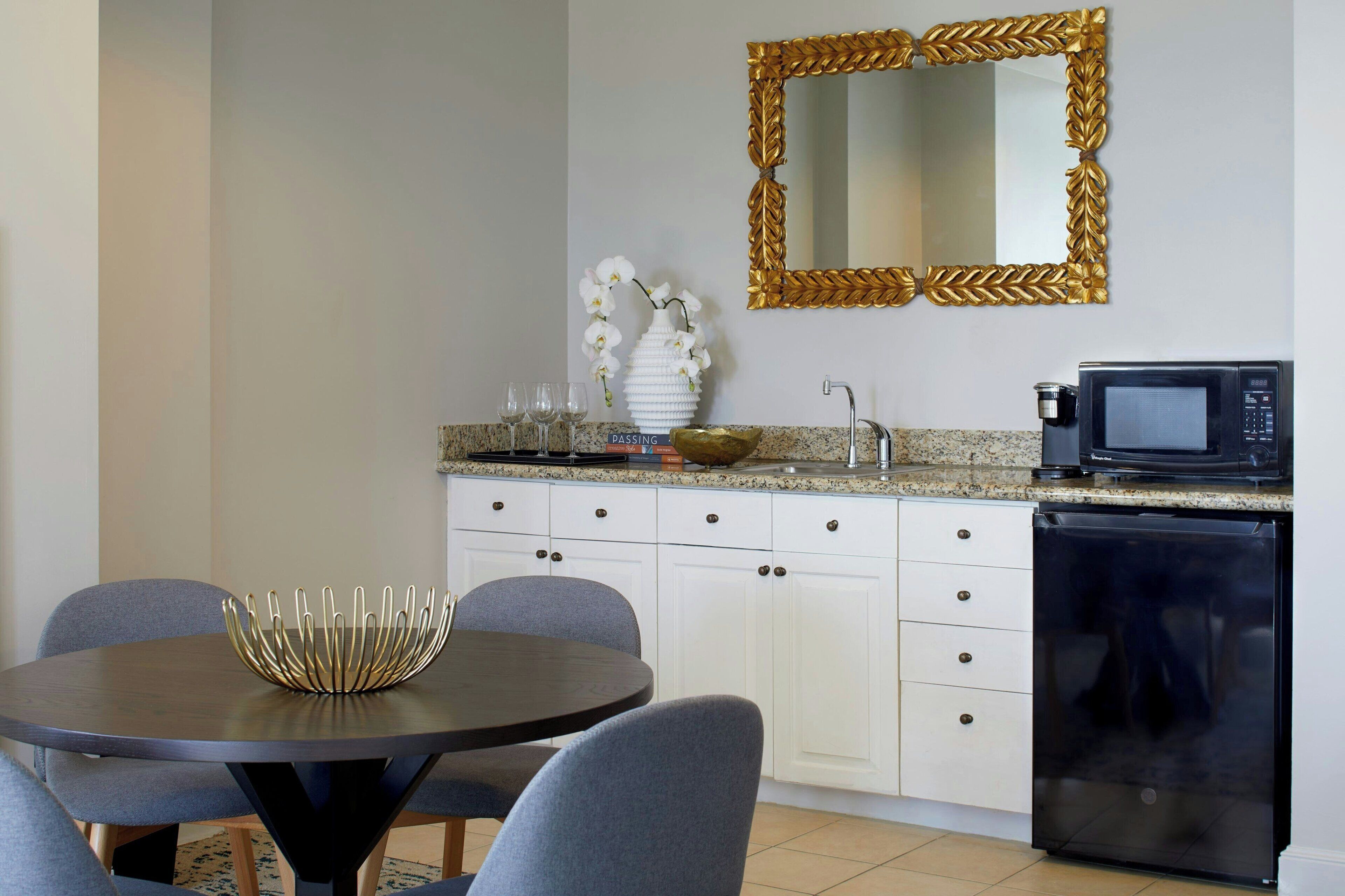 A compact kitchen with white cabinets, a granite countertop, a gold-framed mirror, a black microwave, and a small dishwasher.