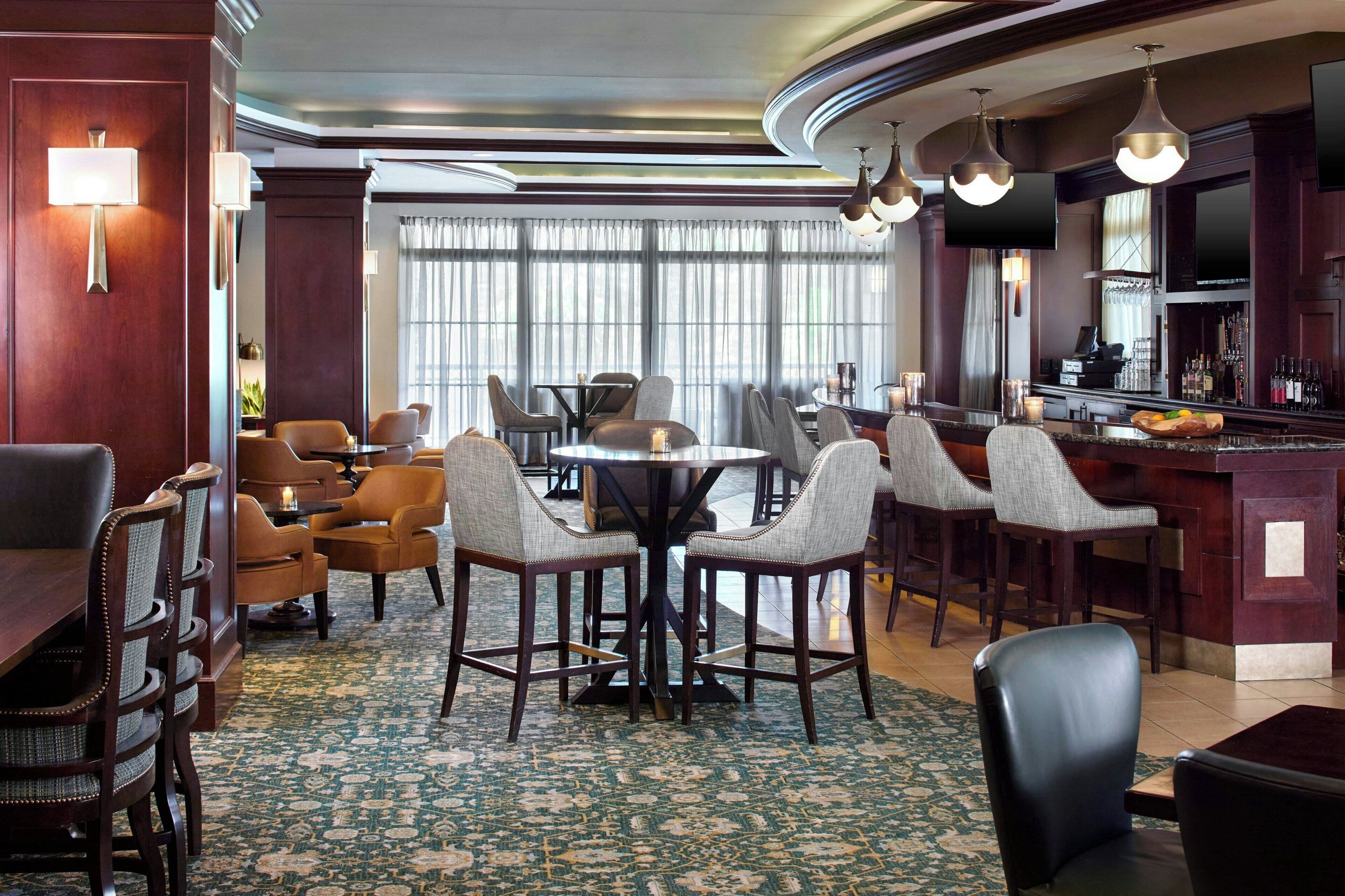 A hotel lobby with a round dining table surrounded by chairs, a bar area with a counter and bar stools, and a large window with curtains.