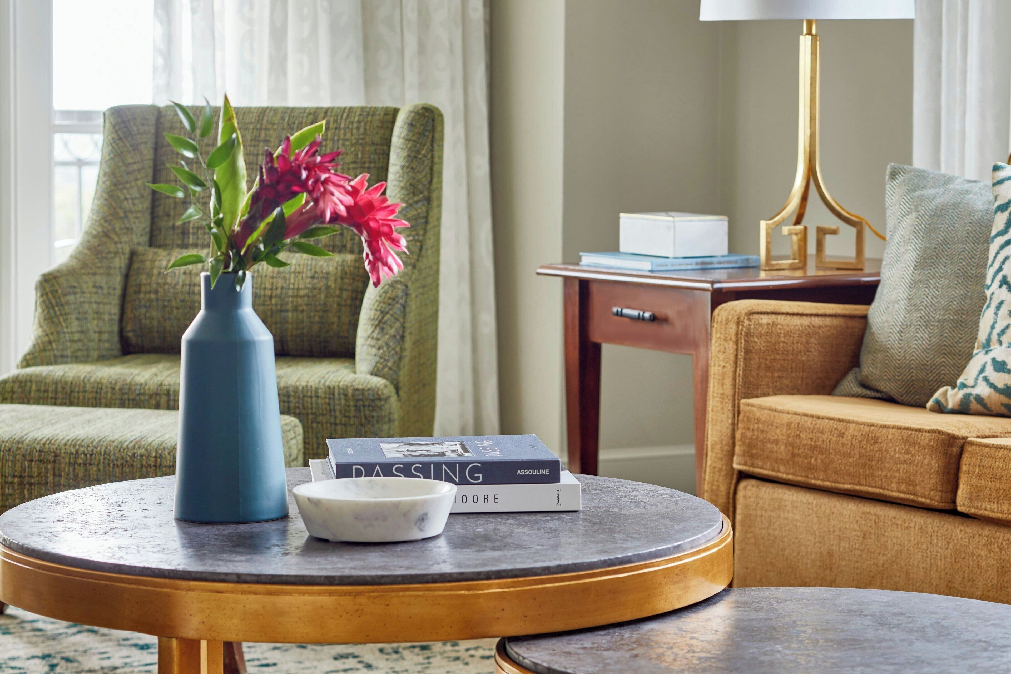 A round table with a vase of flowers, a bowl, and books. A green armchair and a brown sofa are in the background.