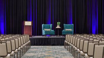 A conference room with rows of chairs facing a stage, a podium, and two armchairs with a small table between them.