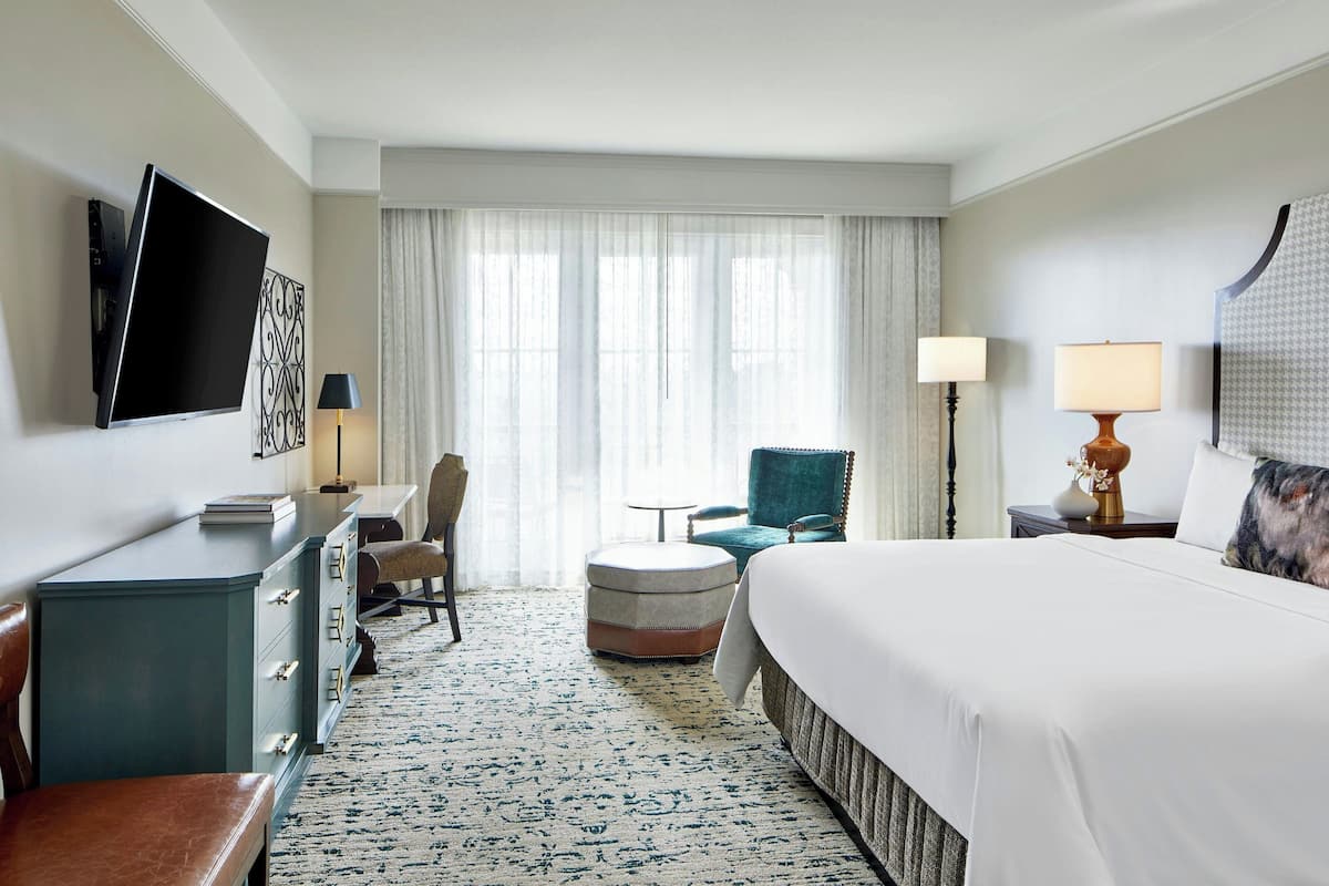 A hotel room with a large bed, a desk with a flat-screen TV, a chair, a small table, and a lamp.