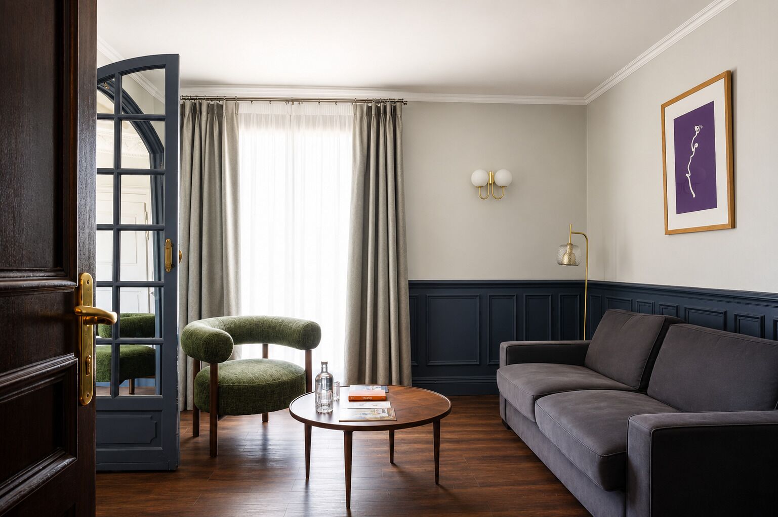 A room with a dark wood door, a green upholstered chair, a grey sofa, a round wooden coffee table, and a framed artwork on the wall.