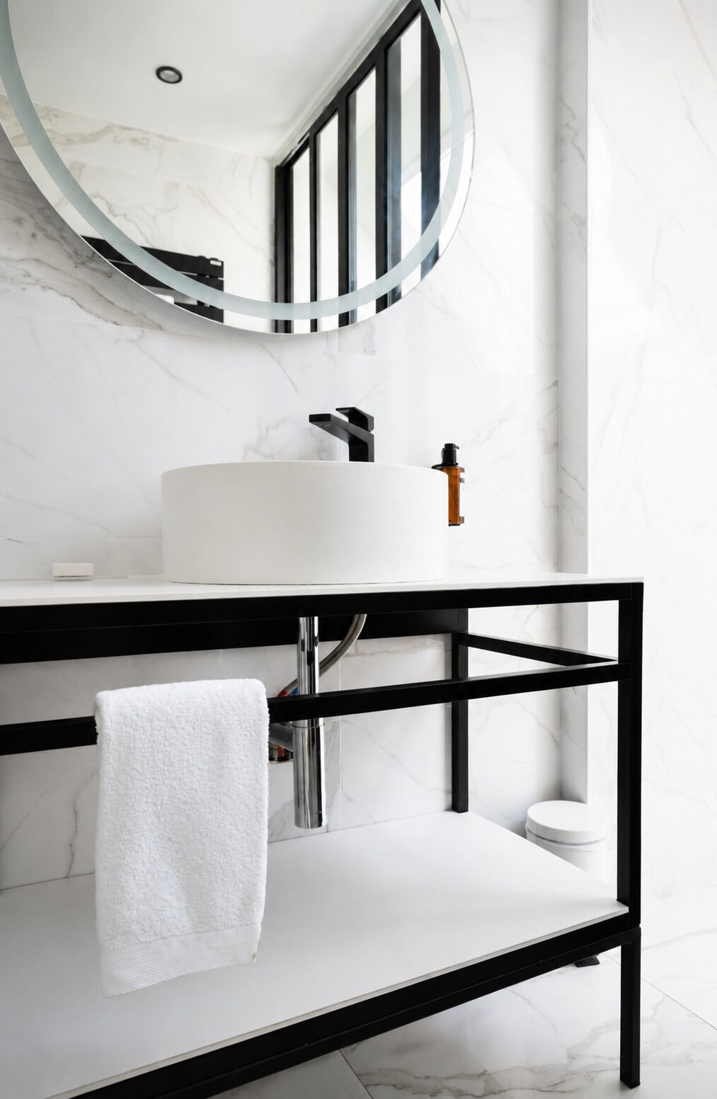A modern bathroom with a white marble sink, a black metal vanity, a white towel, and a round mirror.