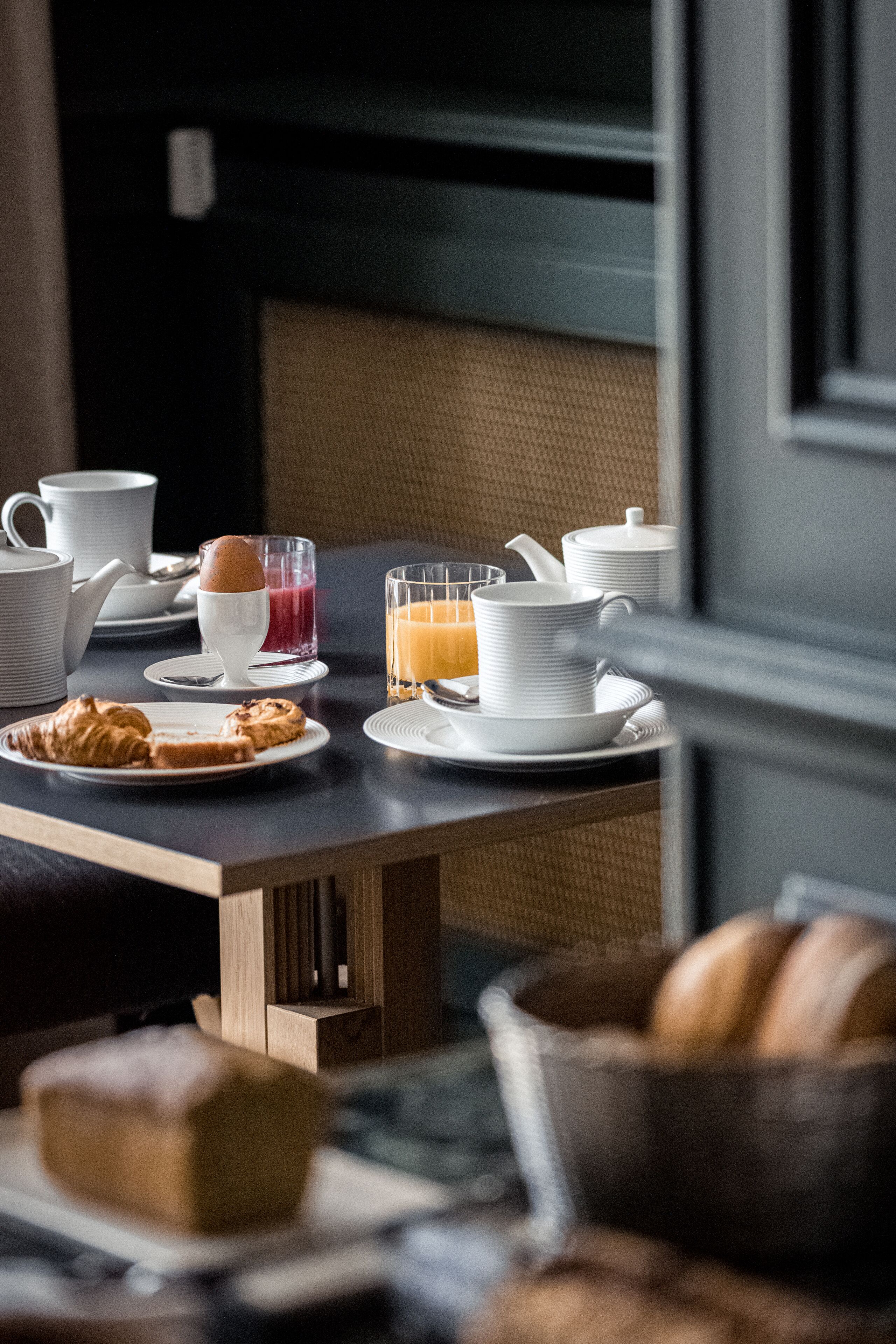 A breakfast setting with a table set for two, including plates, cups, a teapot, and a glass of orange juice.