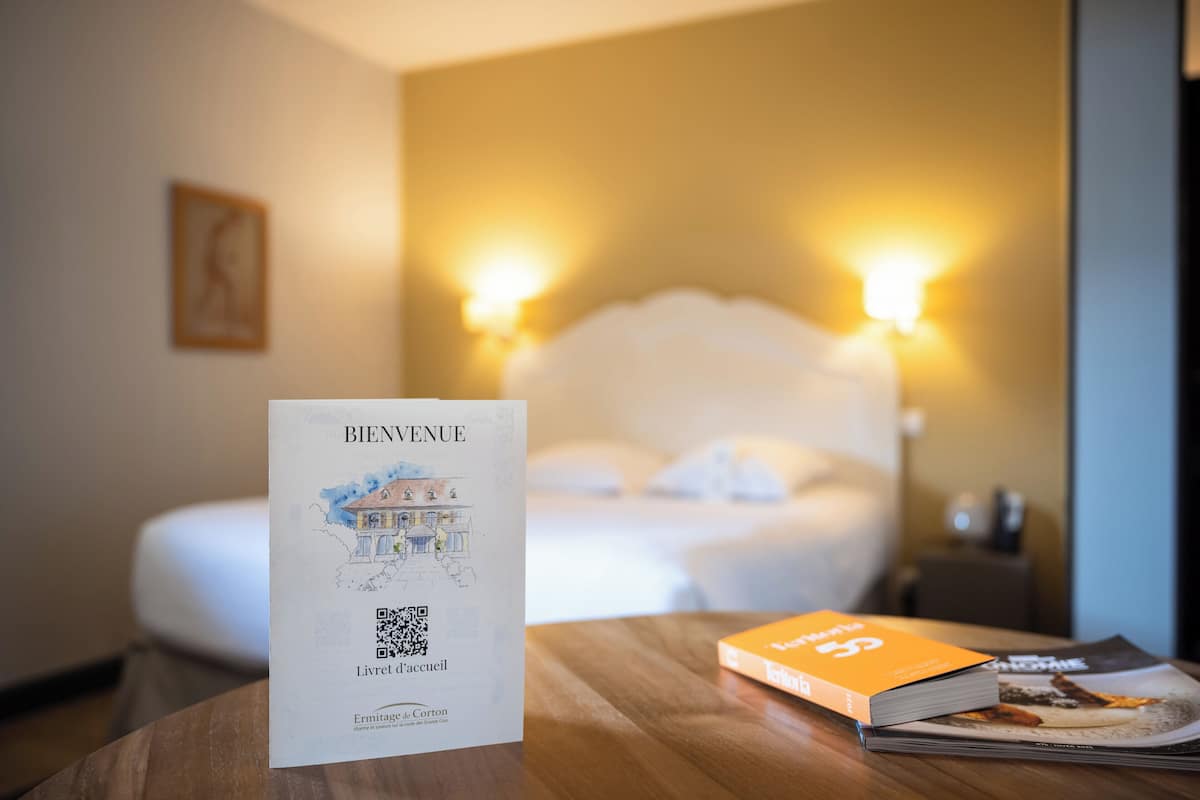 Una habitación de hotel con una cama, una mesita de noche con un libro y una tarjeta de bienvenida, y dos lámparas de pared.