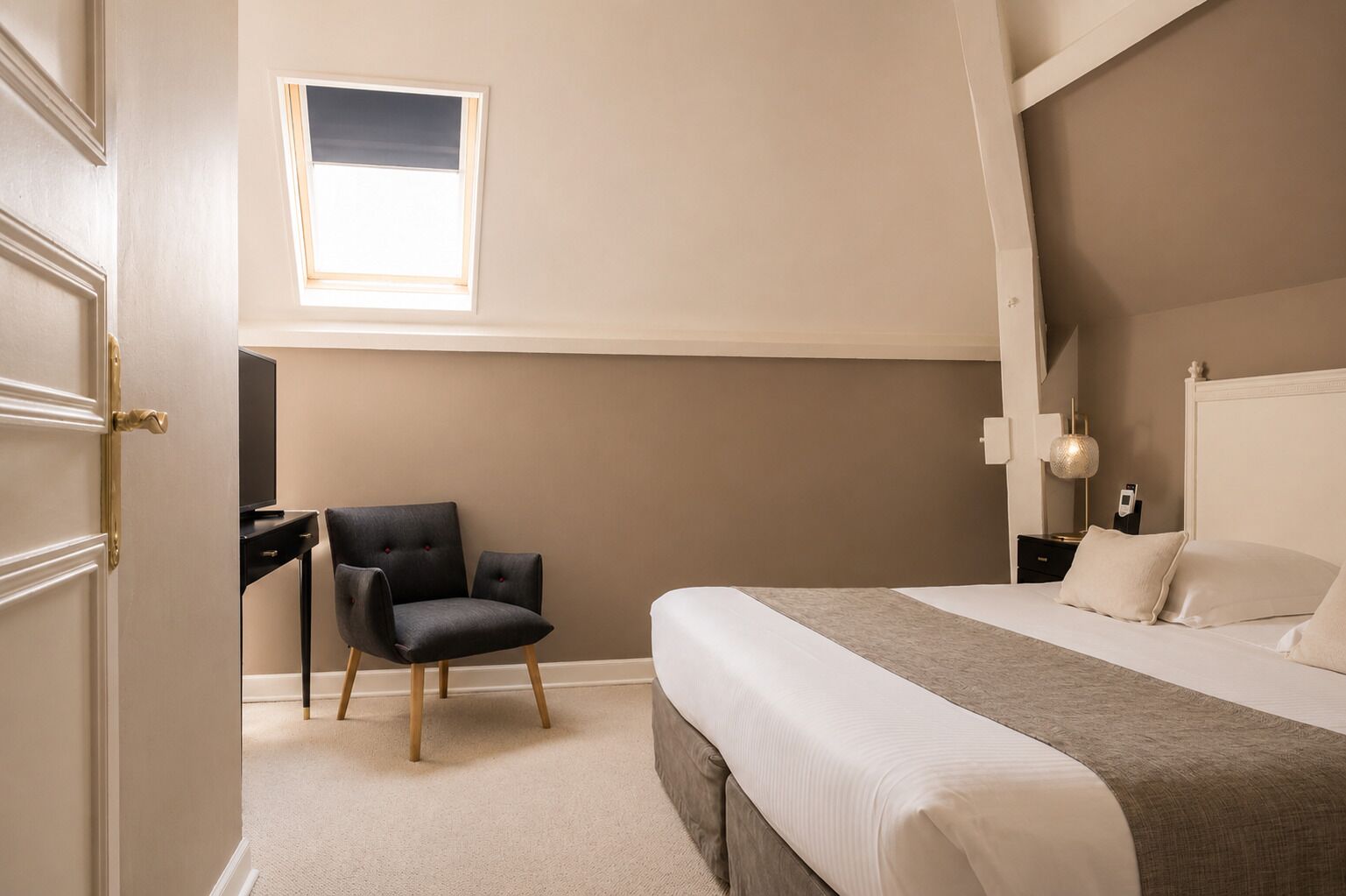 Habitación de hotel con cama, escritorio, silla, ventana y mesita de noche con lámpara.