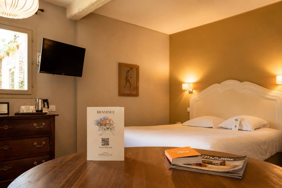 Ein Hotelzimmer mit einem Bett, Nachttischlampen, einem Fernseher, einer Holzkommode, einem Willkommensschild und einem Tisch mit einer Zeitschrift.