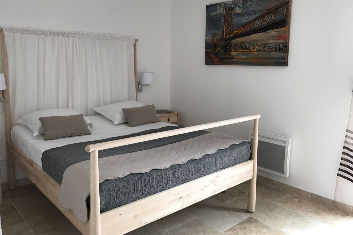 A neatly arranged bedroom with a bed, a nightstand, a lamp, and a painting of a bridge on the wall.