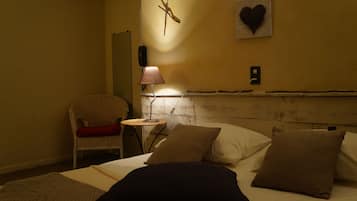 Quarto de hotel com cama, almofadas, uma cadeira, uma mesinha, abajur, relógio de parede e uma decoração em formato de coração.