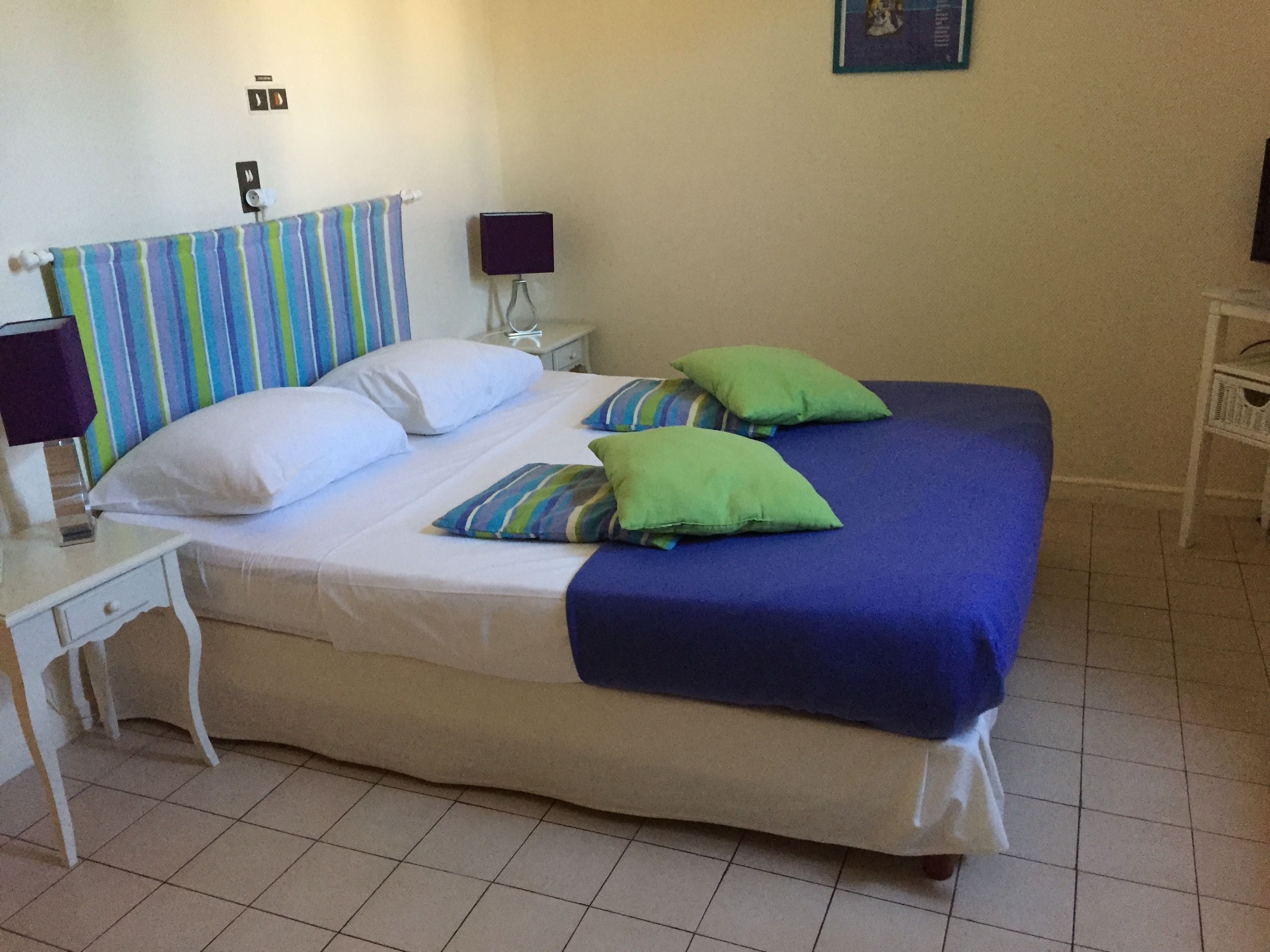 A bed with a striped headboard, two green pillows, and a blue blanket. There is a white nightstand with a lamp and a small table with a lamp. The room has tiled flooring and a wall-mounted light fixture.