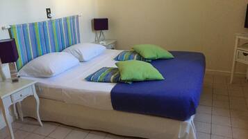 A bed with a striped headboard, two green pillows, and a blue blanket. There is a white nightstand with a lamp and a small table with a lamp. The room has tiled flooring and a wall-mounted light fixture.