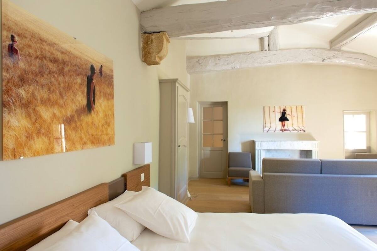 A hotel room with a bed, a large painting, a wooden ceiling, a sofa, and a small table with a lamp.