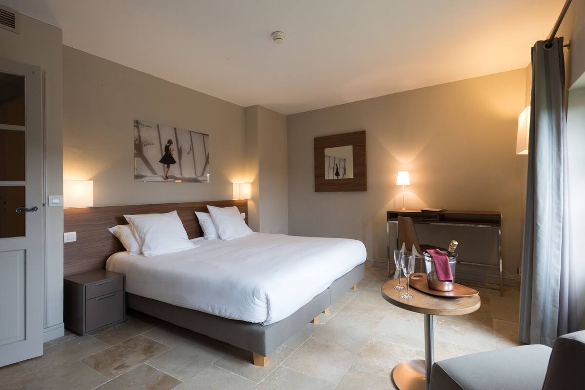 A modern hotel room with a large bed, bedside tables, a desk with a chair, and artwork on the walls.