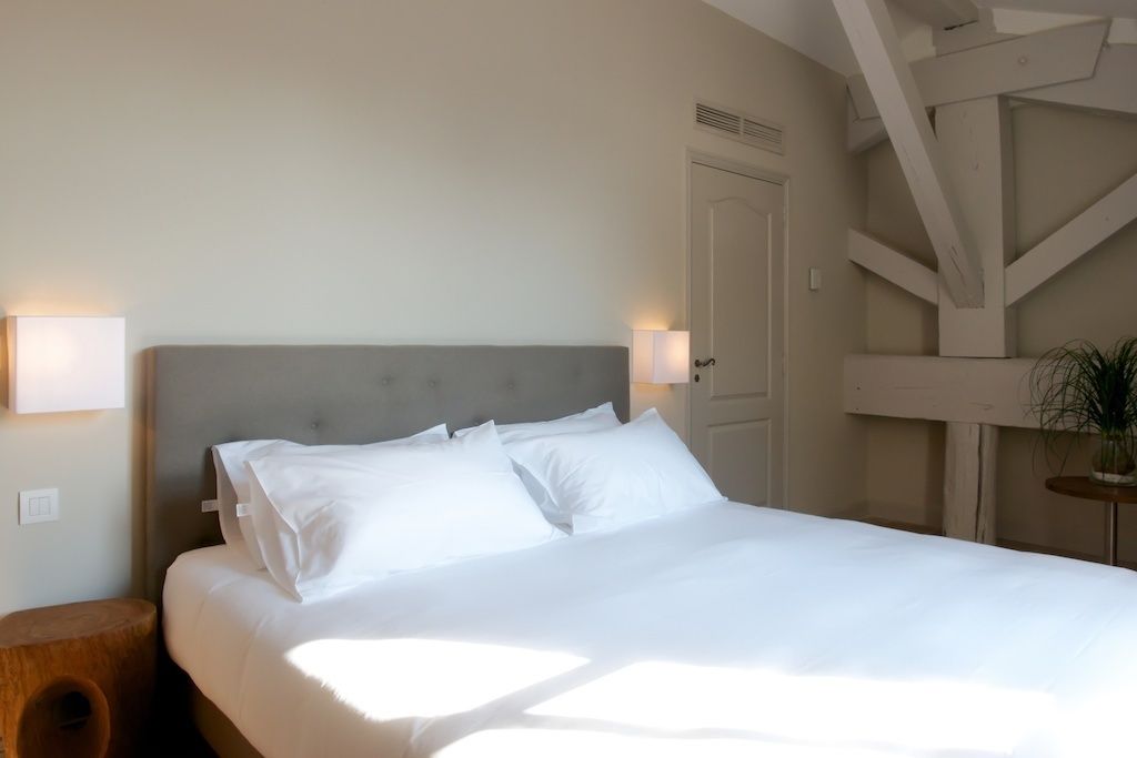 A neatly made bed with white linens, a wooden nightstand, and a wall-mounted light fixture.