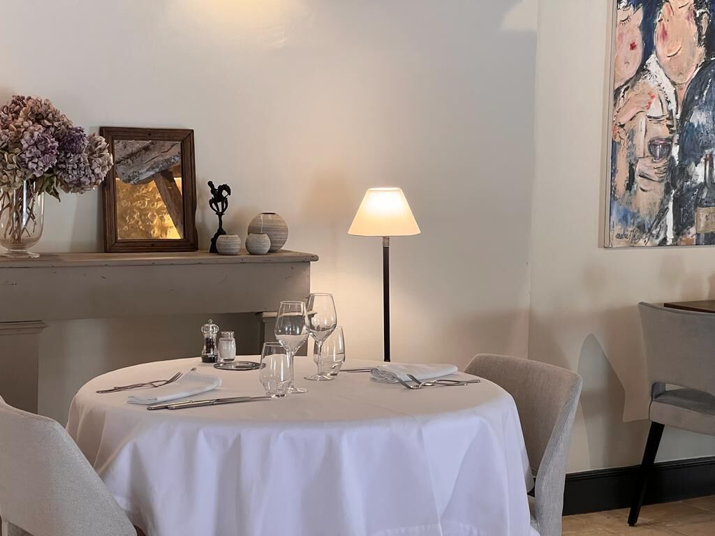Un comedor con una mesa redonda dispuesta para dos personas, un florero, un cuadro enmarcado, una lámpara y un cuadro en la pared.